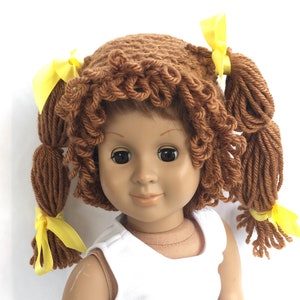 May include: A brown yarn doll wig with two yellow bows. The wig is styled in a curly, messy bun with two pigtails.