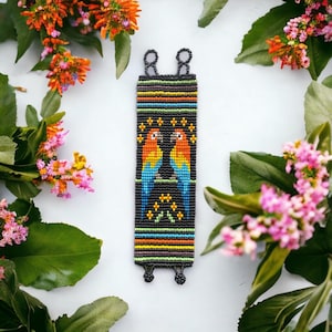 May include: A colorful beaded bracelet with a design of two parrots facing each other. The bracelet is made with black, orange, blue, green, and yellow beads. The background is white.