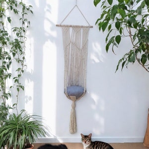 Op de afbeelding: Een witte macramé kattenhangmat met een grijs kussen dat aan het plafond hangt. Een kat zit op de vloer voor de hangmat.