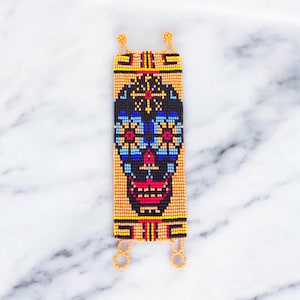 Puede incluir: Una pulsera de cuentas con un diseño de calavera de azúcar. La calavera es negra con detalles azules, rojos y amarillos, sobre un fondo amarillo. La pulsera tiene lazos naranjas en cada extremo.