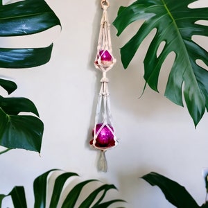 May include: A white macrame plant hanger with two pink glass ornaments hanging from it. The hanger is suspended from a white ceiling.