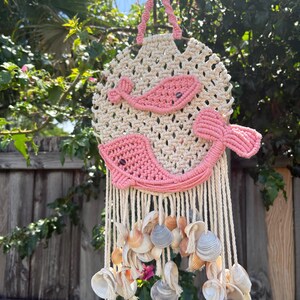 May include: Macrame wall hanging featuring a cream-colored circular design with pink whale accents. The bottom of the wall hanging is adorned with seashells and cream-colored tassels. The wall hanging is suspended by a pink macrame loop.
