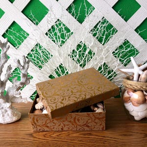 May include: A gold decorative box with a floral pattern is open and filled with seashells. A glass jar filled with seashells is next to the box. A white coral branch is on the left side of the image.