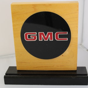 May include: A wooden plaque with a black circle and the word "GMC" in red letters on a silver background.