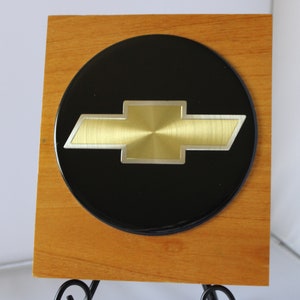 May include: A gold Chevrolet bowtie logo on a black circular plaque mounted on a wooden plaque.