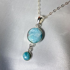 Puede incluir: Un collar de cadena de plata con un colgante que presenta dos piedras redondas de color azul verdoso. La piedra más grande está en la parte inferior del colgante y la más pequeña en la parte superior.