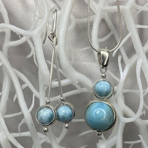 May include: A silver necklace and earrings set with blue gemstone pendants. The necklace features a larger round pendant with a smaller round pendant above it. The earrings have a single round pendant on each earring.