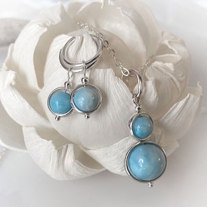 Può includere: Un set di gioielli composto da un paio di orecchini a cerchio in argento e una collana con pendente abbinata, entrambi caratterizzati da pietre rotonde color azzurro lucido incastonate in cornici d'argento. Il pendente presenta due pietre diverse.
