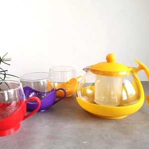 May include: A clear glass teapot with a yellow plastic base, handle, lid, and internal strainer, accompanied by three glass cups featuring red, purple, and orange plastic bases and handles, arranged in a group on a flat surface.