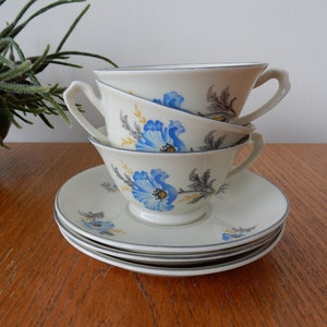 Op de afbeelding: Een set van drie witte theekopjes met blauwe en gele bloemenmotieven. De kopjes zijn op elkaar gestapeld en staan op een houten tafel.