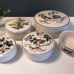 Op de afbeelding: Set van vijf witte, ronde keramische doosjes met bloemmotieven. De doosjes tonen botanische illustraties van verschillende planten en bloemen, waaronder blauwe bessen, cranberry's en gele bloemen. De doosjes zijn allemaal rond en hebben een licht verhoogde rand.