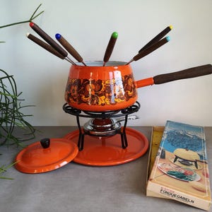 Op de afbeelding: Een oranje fonduepan met een bloemmotief, een zwarte metalen standaard en een bijpassende deksel. De pan heeft een houten handvat en wordt vergezeld door fonduevorken met gekleurde uiteinden. Een doos met de tekst "FONDU GABELN" is ook aanwezig.