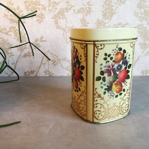 Könnte beinhalten: Eine vintage Creme-farbene Dose mit einem Blumendesign mit roten, gelben und violetten Blumen. Die Dose hat einen goldenen Rand und ein dekoratives Muster.