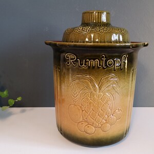 May include: A brown and green ceramic jar with a lid. The jar has a raised design of fruit and the word "Rumtopf" on the front.