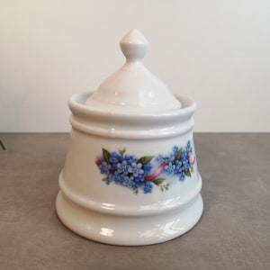 Op de afbeelding: Witte porseleinen suikerpot met deksel, versierd met een bloemmotief van blauwe vergeet-me-nietjes en een roze lint.