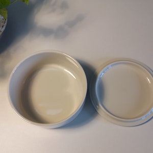 Może przedstawiać: Biała ceramiczna miska z dopasowaną pokrywką. Miska jest okrągła i ma gładkie, błyszczące wykończenie.