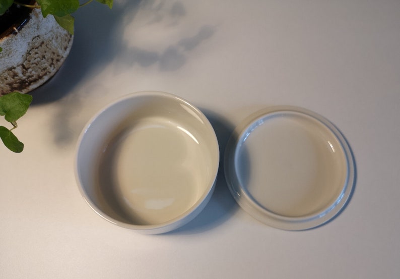 Może przedstawiać: Biała ceramiczna miska z dopasowaną pokrywką. Miska jest okrągła i ma gładkie, błyszczące wykończenie.