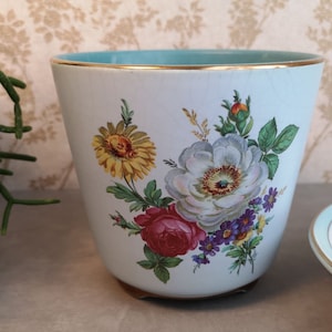 Op de afbeelding: Een witte keramische pot met een blauwe rand en gouden rand. De pot heeft een bloemenmotief met witte, rode, gele en paarse bloemen.