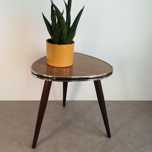 Op de afbeelding: Een driepotige bijzettafel in mid-century modern stijl met drie donkere houten poten en een messing rand. Het tafelblad is van houtlaminaat met een bruin houtnerfpatroon. Een gele keramische pot met een groene succulent staat op de tafel.