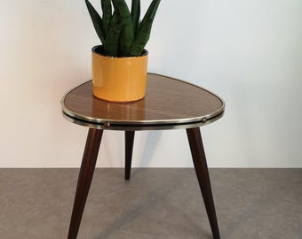 Może przedstawiać: Trójkątny stolik pomocniczy w stylu mid-century modern z trzema ciemnymi drewnianymi nogami i mosiężną obwódką. Blat stołu wykonany jest z laminatu drewnopodobnego z brązowym wzorem słojów drewna. Na stole stoi żółty ceramiczny pojemnik z zieloną rośliną sukulent.