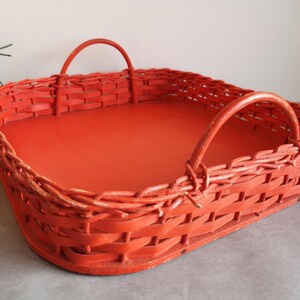 Op de afbeelding: Een vierkante, oranje rieten mand met twee gebogen handvatten. De mand heeft een effen oranje bodem en is gemaakt van geweven riet. De mand is leeg en staat op een grijze ondergrond.