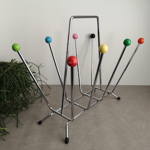 Pode incluir: Um porta-revistas de cromo com seis pontas em forma de bola coloridas nos braços. O porta-revistas tem uma base retangular e uma moldura retangular.