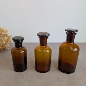 Op de afbeelding: Drie apothekersflessen van amberkleurig glas in verschillende hoogtes, elk met een cilindrisch lichaam, een smalle hals en een bijpassende donkerbruine glazen stop. De flessen staan op een rij en hebben een warme bruine tint.