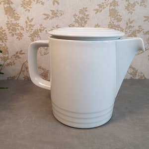 Op de afbeelding: Witte theepot van keramiek met een deksel en een handvat. De theepot heeft een eenvoudig ontwerp met een rond lichaam en een tuit.