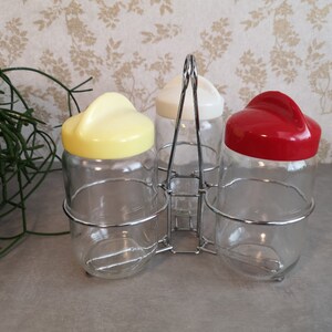 Op de afbeelding: Set van drie glazen kruidenpotten met gekleurde plastic deksels in geel, wit en rood. De potten worden bij elkaar gehouden door een verchroomd draadrek.