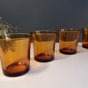 Op de afbeelding: Vier amberkleurige glazen tumblers op een witte ondergrond. De glazen zijn leeg en hebben een ronde bodem.