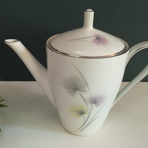Op de afbeelding: Witte theepot van keramiek met een deksel en een bloemenmotief. De theepot heeft een zilveren rand rond het deksel en een zilveren lijn rond de basis. Het bloemenmotief heeft roze, gele en grijze bloemen.