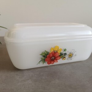 Op de afbeelding: Witte rechthoekige container met een deksel, versierd met een bloemenontwerp met rode, gele en witte bloemen. De container is waarschijnlijk gemaakt van glas of keramiek en is geschikt voor het bewaren van voedsel.