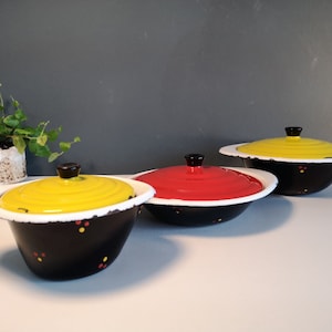 Op de afbeelding: Drie vintage emaille kommen met deksels. De kommen zijn zwart met witte randen en gele, rode en zwarte stippen. De deksels zijn geel, rood en wit.