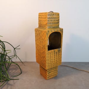 Op de afbeelding: Een hoog, rechthoekig, geel en bruin decoratief object met een getextureerd oppervlak en een boogvormige opening. Het object heeft een vierkante basis en een lus aan de bovenkant. Een dun, bruin snoer komt uit de basis.