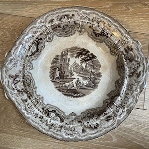 Peut inclure: Assiette ancienne en céramique marron et blanche, ornée d'un paysage détaillé au centre. Le bord de l'assiette est festonné avec un motif répétitif de bâtiments et de feuillage. Une pièce décorative vintage.