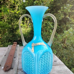 Op de afbeelding: Een turquoise glazen vaas met twee heldere, gedraaide handvatten. De vaas heeft een vierkante basis met een geweven patroon en een uitlopende bovenkant. Een gouden label leest "palina fiorentina".