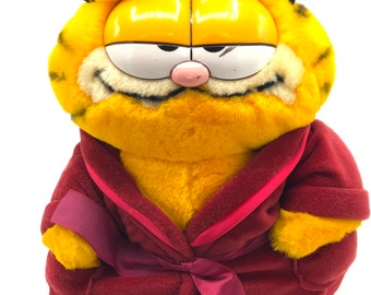 May include: A plush Garfield toy wearing a maroon robe with a matching sash. The toy has a yellow and orange striped body, a white face, and black whiskers.
