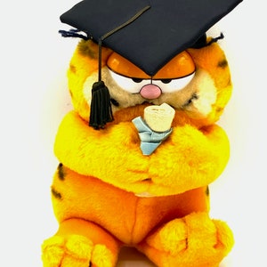 May include: A plush orange and black Garfield cat wearing a black graduation cap and holding a blue diploma.