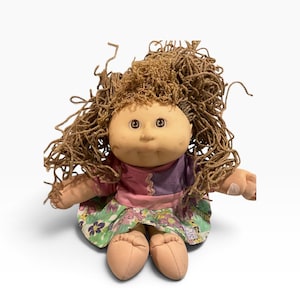 May include: A soft-sculpture doll with light skin, brown yarn hair, and brown eyes. The doll wears a short-sleeved top with purple and pink panels and a skirt featuring a green, purple, and pink floral pattern. The doll has visible stitched toes.