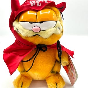 May include: A plush orange Garfield cat wearing a red devil costume with horns and a cape. The costume has the word "DEVIL" written on the front. The cat is holding a black chain in its paw. The plush toy has a tag attached to it.