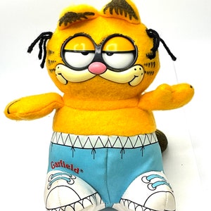 May include: A plush Garfield toy wearing blue shorts and white sneakers. The toy has a yellow body, black eyes, and a red mouth. The text "Garfield" is printed on the shorts.