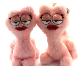 May include: Two plush pink cat-like creatures with exaggerated features. They have large, sleepy-looking eyes, red lips, and black eyelashes. The toys are soft and fluffy, with a whimsical and slightly unsettling appearance.