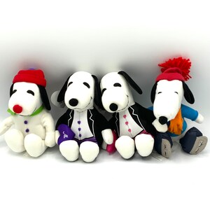 May include: Four plush Snoopy toys in different outfits. One is wearing a red and white Santa hat, another is wearing a black tuxedo, another is wearing a pink dress, and the last one is wearing a blue and white winter outfit with a scarf.