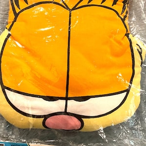 Peut inclure: Un gant de cuisine orange sur le thème de Garfield, avec des contours noirs et un visage jaune. Le gant est emballé dans du plastique transparent. Une étiquette en carton assortie indique "Garfield Cooking Mitt".