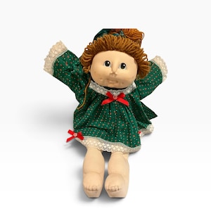 Peut inclure: Une poupée en tissu avec des cheveux en laine brune et un visage rond. La poupée porte une robe vert foncé avec un petit motif floral rouge, une bordure en dentelle blanche aux manches et à l'ourlet, et deux petits nœuds rouges sur le devant.