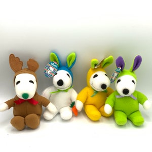 May include: Four plush toys dressed in different costumes. The first is a brown reindeer, the second is a white bunny with blue ears, the third is a yellow bunny, and the fourth is a green bunny with purple ears.