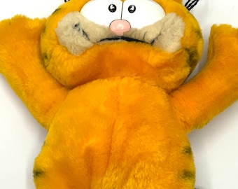 Puede incluir: Un peluche de Garfield de color naranja brillante con rayas negras y una nariz rosa. El juguete tiene ojos blancos grandes y está en una pose juguetona con los brazos levantados. El peluche está hecho de material suave y difuso.