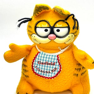 May include: A plush Garfield toy in bright orange with black accents. The toy has large, expressive eyes, a pink nose, and a smiling mouth. It wears a bib with a blue and white checkered pattern and the words "Garfield LASAGNA BIB".