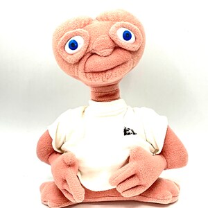 May include: A plush toy of the alien character E.T. from the movie E.T. the Extra-Terrestrial. The toy is wearing a white t-shirt with a black logo on the front. The toy is sitting with its arms crossed and has a friendly expression on its face.