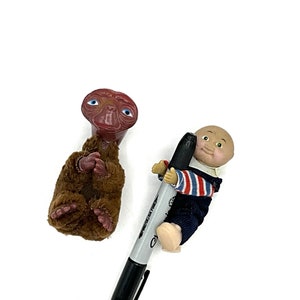 May include: An E.T. figurine and a doll are featured in this image. The E.T. figurine is brown with large eyes. The doll is wearing a blue and white striped shirt and blue overalls. A black Sharpie pen is also in the image.